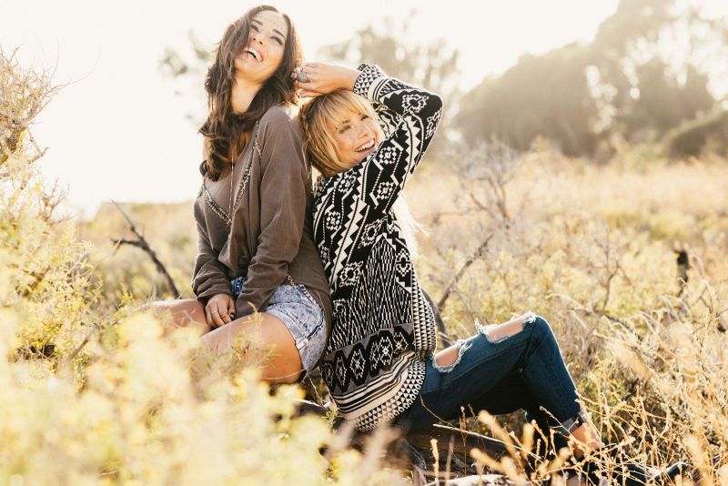 Bohemian lifestyle shot of two friends laughing together in a sunny natural landscape. The image highlights fall-inspired fashion, including a geometric knit cardigan, an embroidered tunic, and ripped denim.