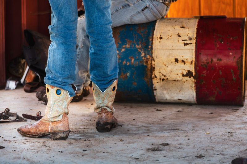 detail shot of a person wearing intricately stitched cowboy boots standing next to someone sitting on a faded red, white, and blue rodeo barrel in Willits, California.