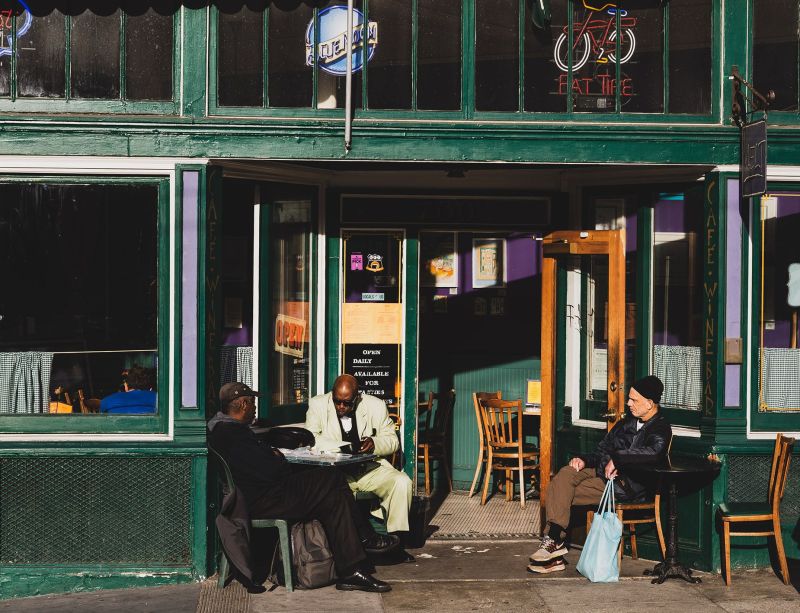 Three men relaxing at outdoor tables in front of a classic green-painted cafe storefront in bright sunlight on Columbus Ave, San Francisco.
