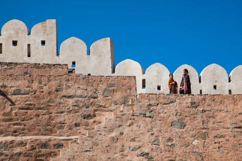 Two people wrapped in traditional shawls standing atop the massive, ancient stone walls of Kumbhalgarh Fort in Rajasthan, India, featuring its distinctive rounded parapets against a clear blue sky.