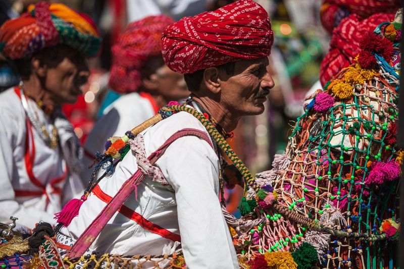 An Indian man wearing a bright red turban and white shirt, surrounded by vibrant, multicolored tasseled harnesses at a cultural festival.