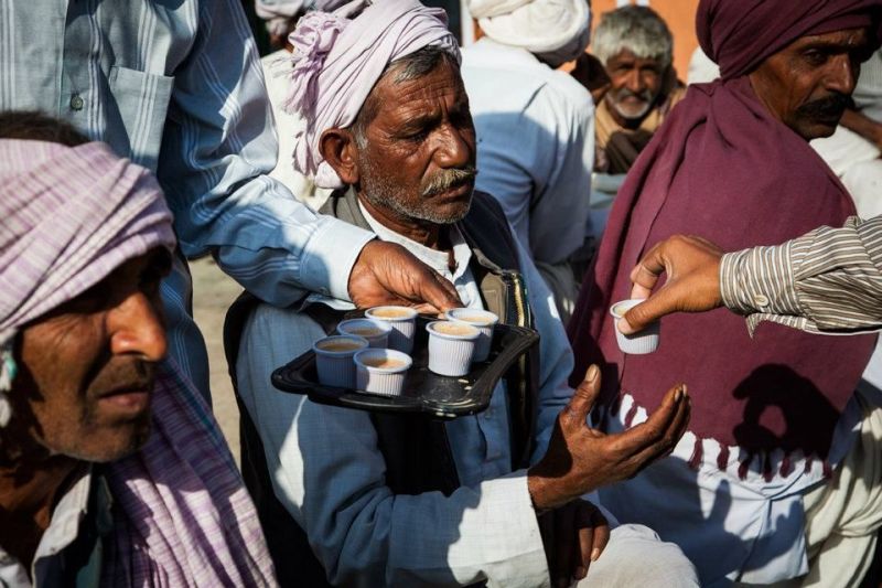 A candid, close-up shot of several older men in traditional Indian attire, including turbans and shawls, gathered together in bright sunlight. In the center, a hand holds a black tray carrying several small white cups filled with milky tea, as another per